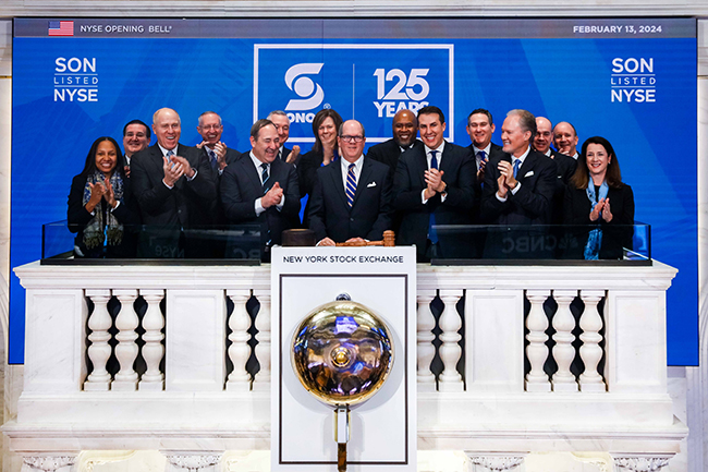 Group of business people wearing suits smiling and clapping in front of a wall that reads"SON LISTED NYSE, SONOCO 125 Years" Standing in front of the New York Stock Exchange bell.